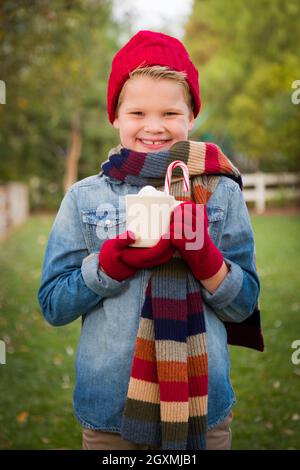 Hübscher junger Junge trägt Weihnachtskleidung, hält heißen Kakao mit Marshallows und Candy Cane draußen. Stockfoto