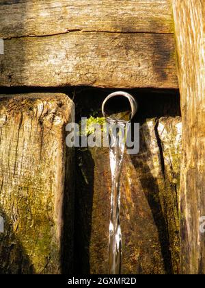 Natürliche Dorfquelle mit sauberem Trinkwasser. Wasser fließt aus einem Eisenrohr aus einer Holzwand. Reines Wasser, das aus dem Rohr fließt und fällt Stockfoto