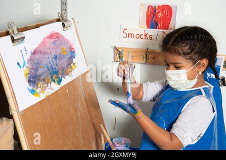Bildung Vorschule 4-5-jähriges Mädchen in der Smck Malerei an Staffelei, Pinsel Farbe auf ihre eigene Hand, trägt Gesichtsmaske, um gegen Covid-19 zu schützen Stockfoto