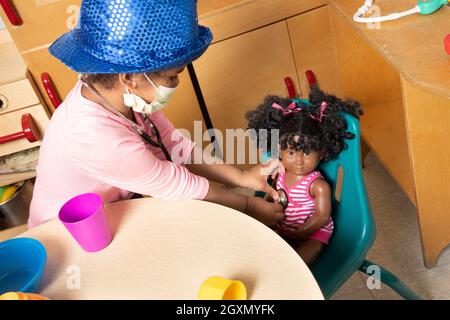 Bildung Vorschule 3-4-Jährige geben vor, im Familienbereich zu spielen, Mädchen benutzen Stethoskop auf der Puppe, tragen Spielhut & Gesichtsmaske gegen Covid-19-Virus Stockfoto