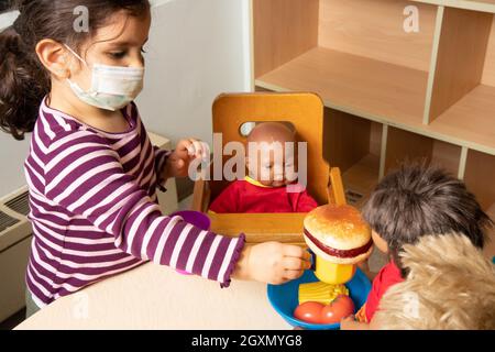 Bildung Vorschule 3-4-Jährige geben vor, im Familienbereich zu spielen, Mädchen füttern Puppe mit Spielfutter, tragen Gesichtsmaske, um gegen Covid-19i zu schützen Stockfoto