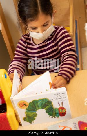 Bildung Vorschule 3-4-jähriges Mädchen, das sich das Buch Eric Carle's The Very Hungry Caterpillar mit Gesichtsmaske anschaut Stockfoto