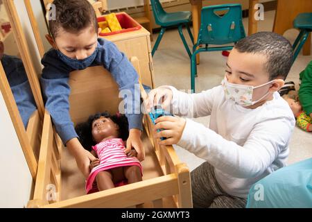 Bildung Vorschule 3-4-jährige zwei Jungen spielen im Familienbereich mit Puppe und Puppenbett, einer spielt mit Spielzeugeisenbahn, trägt Gesichtsmasken zum Schutz Covid Stockfoto