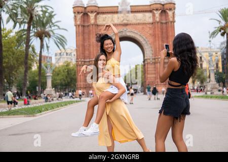 Unbeschwerte junge multirassische Freundinnen, die gemeinsam Sommerferien genießen und lustige Fotos gegen den Triumphbogen während einer Tour in Barcelona City machen Stockfoto