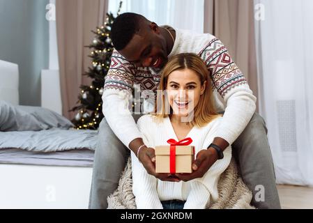 Eine junge kaukasische Frau erhält ein Geschenk von ihrem schwarzen Ehemann. Romantisches Paar Austausch Weihnachtsgeschenke Zu Hause. Stockfoto