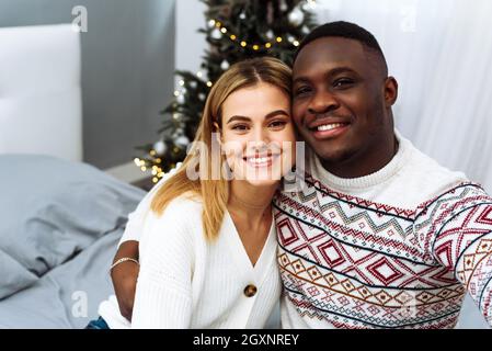 Ein schönes multiethnisches Paar umarmt sich zu Hause vor dem Hintergrund eines Weihnachtsbaums und nimmt ein Selfie auf. Einen glücklichen Moment festhalten. Stockfoto