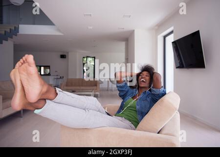 junge schöne afroamerikanische Frau hört gerne Musik mit Kopfhörern und Tablet in Ihrem Sessel in ihrem Luxushaus Stockfoto