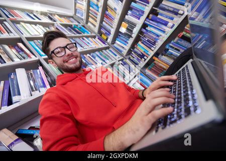 Ein junger Student mit Brille sitzt in der Bibliothek, liest ein Buch und macht sich mit einem Latpop Notizen Stockfoto