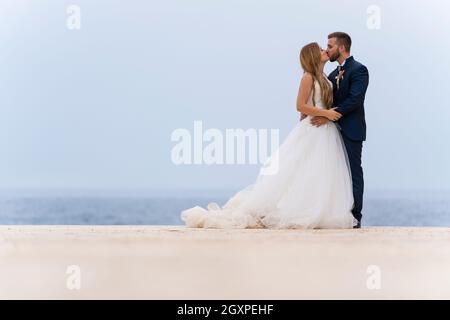 Frisch verheiratetes Paar küsst und umarmt sich mit dem Meer im Hintergrund Stockfoto