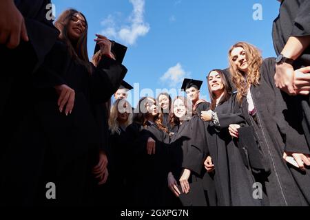 Herzlichen Glückwunsch! Low-Angle-Ansicht der glücklichen Gruppe von sechs jungen fröhlichen Absolventen in schwarzen Kleidern, werfen ihren Kopf tragen in der Luft und feiern, Stockfoto