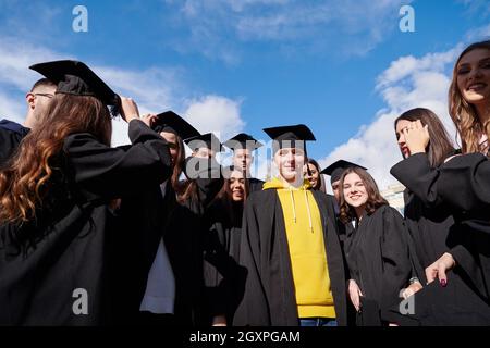 Herzlichen Glückwunsch! Low-Angle-Ansicht der glücklichen Gruppe von sechs jungen fröhlichen Absolventen in schwarzen Kleidern, werfen ihren Kopf tragen in der Luft und feiern, Stockfoto