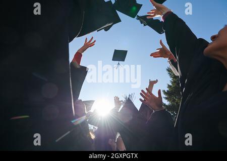 Herzlichen Glückwunsch! Low-Angle-Ansicht der glücklichen Gruppe von sechs jungen fröhlichen Absolventen in schwarzen Kleidern, werfen ihren Kopf tragen in der Luft und feiern, Stockfoto