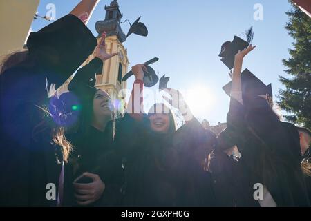 Herzlichen Glückwunsch! Low-Angle-Ansicht der glücklichen Gruppe von sechs jungen fröhlichen Absolventen in schwarzen Kleidern, werfen ihren Kopf tragen in der Luft und feiern, Stockfoto