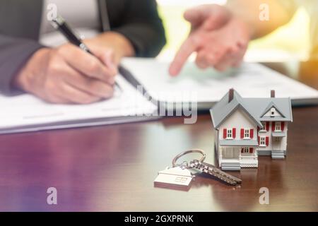 Immobilienmakler und Kunde unterzeichnen Vertragsunterlagen mit Hausschlüsseln und kleinem Modellhaus vor sich. Stockfoto