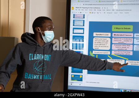 Bildung High School Klassenzimmer männlichen Schüler präsentiert sich der Klasse mit Smart Board, zeigt auf Abschnitt, trägt Gesichtsmaske, um Covid-19 zu schützen Stockfoto