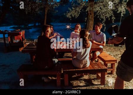 Gruppe von Freunden feiern gerne Urlaub mit Sprinklern und trinken Rotwein beim Picknick französischen Dinner Party im Freien in der Nähe des Ri Stockfoto