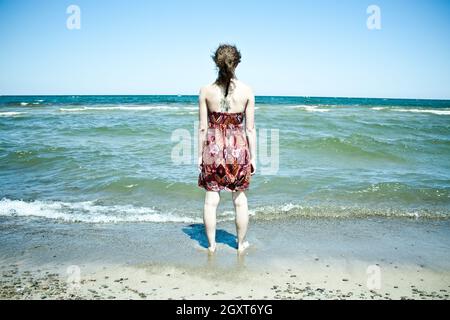 Eine Frau mit langen braunen Haaren, die eine rote Sandress trägt, steht im knöcheltiefen Wasser am Strand, während Wellen an ihre Knöchel schlagen Stockfoto