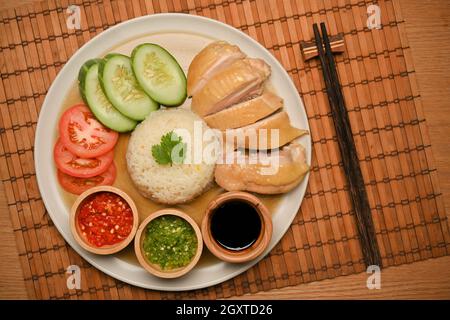 Draufsicht, Nahaufnahme von Hainanese-Hühnerreis mit Gurken- und Tomatenscheiben, Chilisaucen und süßer Sojabohnensauce. Essstäbchen auf dem Tisch. C Stockfoto