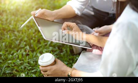 Eine kurze Aufnahme von zwei Freunden, die an einem Sommertag auf einem Tablet über die Arbeit sprechen und auf einem Rasen im Park sitzen Stockfoto