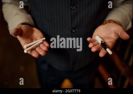Mann hält Zigaretten und Mundstück in den Händen, Bücherregal und reiche Büroeinrichtung auf dem Hintergrund. Tabakrauchkultur, spezifischer Geschmack. Rauch ha Stockfoto