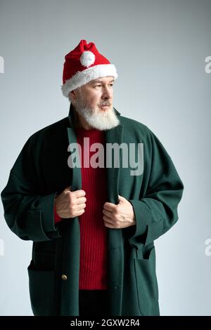 Modischer Mann mittleren Alters mit Weihnachtsmütze, das ein stilvolles Outfit trägt und seinen Mantel anpasst, während er isoliert über hellgrau posiert Stockfoto