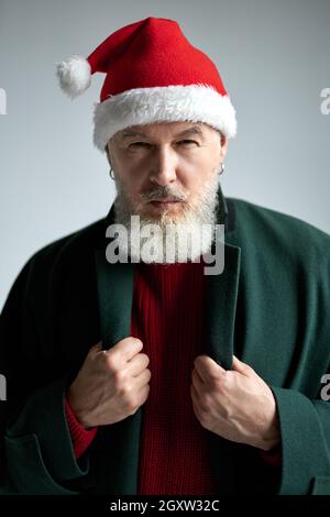 Fokussierter Mann mittleren Alters mit Weihnachtsmütze in stilvollem Outfit, der die Kamera anschaut, seinen Mantel anpasst, während er isoliert über hellgrau posiert Stockfoto