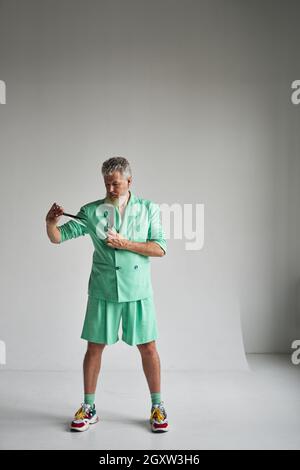 Studio-Aufnahme eines trendigen grauhaarigen reifen Mannes mit Bart, das ein stilvolles Outfit trägt, das konzentriert aussieht, eine scharfe Friseurschere und einen Haarkamm hält und steht Stockfoto