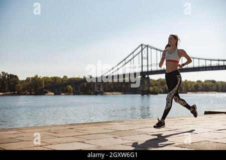 Fröhliche Sportlerin im Trainingsanzug und in Sneakers läuft am Stadtdamm entlang Stockfoto