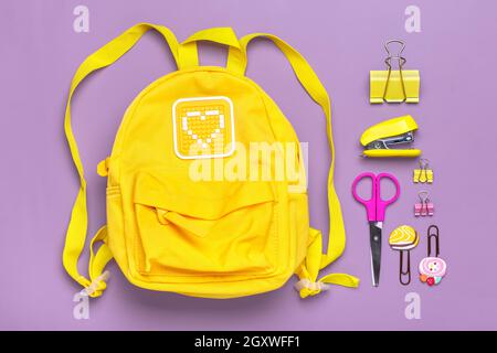 Zurück zur Schule, Bildungskonzept. Gelber Rucksack mit Schulbedarf - Notizbuch, Stifte, Clips, Schere isoliert auf violettem Hintergrund Draufsicht Copy Stockfoto