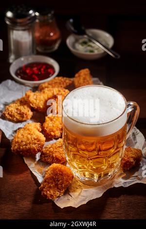 Bier mit Schaum im Glaskrug in der Nähe von gebratenen Hühnchen-Nuggets serviert auf knittertem Papier auf dem Tisch mit Saucen und Gewürzen Stockfoto