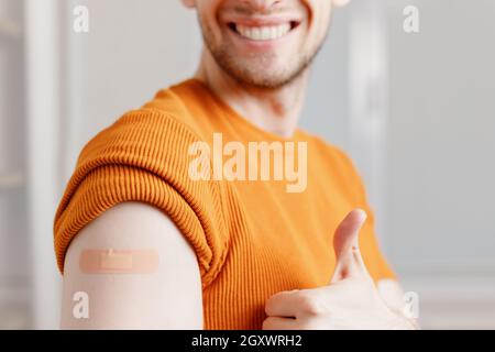 Lächelnder junger Mann mit klebstoffhaftem Verbandsputz auf der Schulter, der nach der Injektionsimpfung den Daumen nach oben zeigt. Nahaufnahme der zugeschnittenen Ansicht Stockfoto