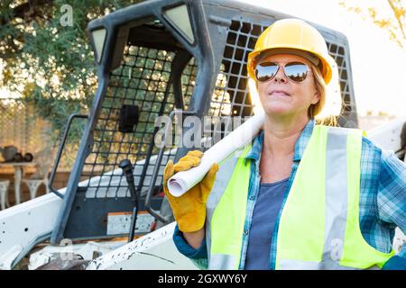 Weibliche Angestellte, Die Technische Blueprints In Der Nähe Des Kleinen Bulldozers Auf Der Baustelle Halten Stockfoto