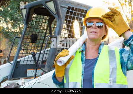 Weibliche Angestellte, Die Technische Blueprints In Der Nähe Des Kleinen Bulldozers Auf Der Baustelle Halten Stockfoto