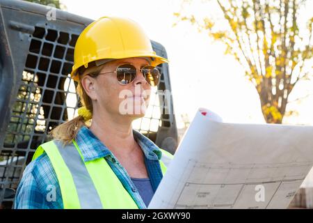 Weibliche Angestellte, Die Technische Blueprints In Der Nähe Des Kleinen Bulldozers Auf Der Baustelle Halten Stockfoto