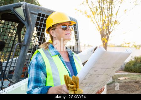 Weibliche Angestellte, Die Technische Blueprints In Der Nähe Des Kleinen Bulldozers Auf Der Baustelle Halten Stockfoto