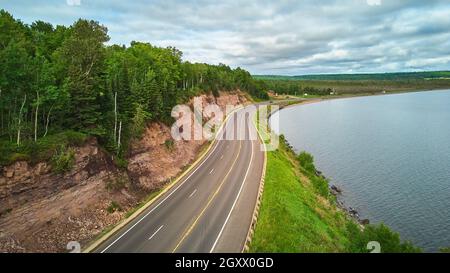 Luftaufnahme über die Straße, die um den See führt, und gegen die felsige Klippe mit grünem Wald Stockfoto