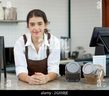 Die jungen kaukasischen Mitarbeiter im Café mit brauner Schürze lehnen sich an die Theke, während sie auf die Kunden warten, die den Laden betreten. Morgenstimmung in einem Stockfoto