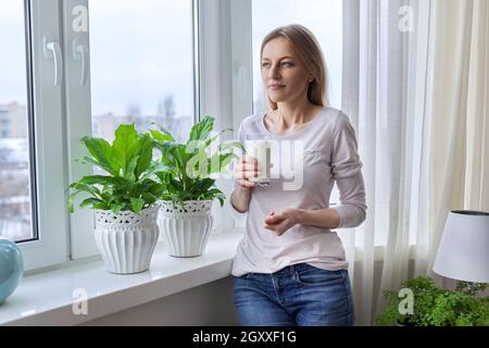 Frau mittleren Alters mit einem Glas Milch, zu Hause am Fenster Stockfoto