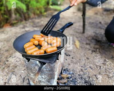 Feuern Sie leckere Würstchen an, die in einer Pfanne über dem Lagerfeuer kochen.Sommerpicknick. Stockfoto