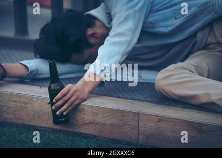 Nach der Party. Betrunkener bärtiger junger Mann, der auf dem Bürgersteig auf der Straße liegt und schläft, nachdem er viel Bier getrunken hat. Stockfoto