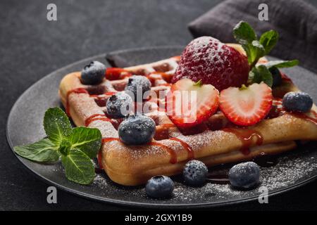 Köstliche Waffeln mit Puderzucker und Beeren auf schwarzem Keramikplatte auf grauem Hintergrund. Süße Mahlzeit. Nachtisch. Serviert werden Speisen Stockfoto