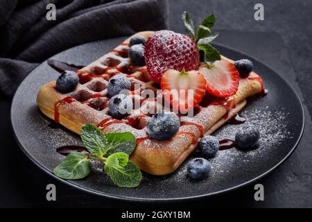 Köstliche Waffeln mit Puderzucker und Beeren auf schwarzem Keramikplatte auf grauem Hintergrund. Süße Mahlzeit. Nachtisch. Serviert werden Speisen Stockfoto