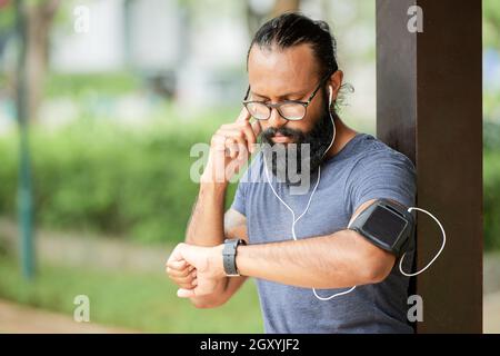 Ernsthafter junger bärtiger Inder in einer Brille, der im Freien steht und Kopfhörer anpasst, während er auf der Smartwatch eine Audiobotschaft hört Stockfoto