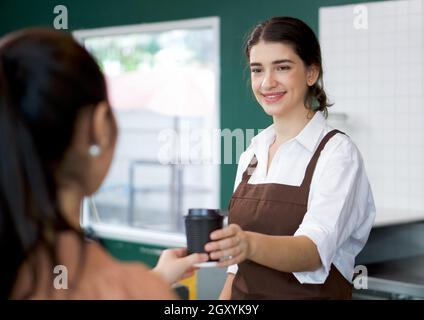 Der kaukasische Barista mit Schürze serviert jungen asiatischen Gästen heißen Kaffee. Morgendliche Atmosphäre in einem Café. Stockfoto