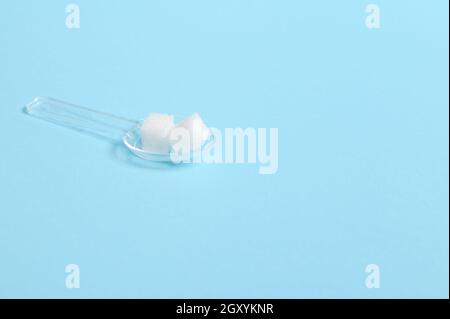 Reine raffinierte weiße Zuckerwürfel auf einem transparenten Messlöffel isoliert auf farbigem blauem Hintergrund mit Kopierfläche für medizinische Konzeptwerbung. Stockfoto