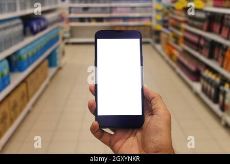 Smartphone mit leerem weißen Bildschirm. Hand hält Smartphone mit verschwommenem Supermarkt Gänge Hintergrund. Online-Shopping-Konzept. Stockfoto