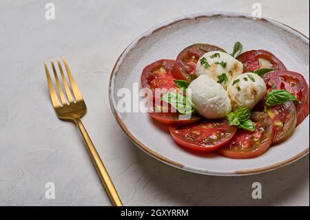 Caprese-Salat mit roten Tomaten, Basilikum und Mozzarella-Käse in weißem Teller und goldener Gabel Stockfoto
