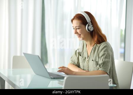 Glückliche Frau mit kabellosen Kopfhörern, die zu Hause ihren Laptop überprüft Stockfoto