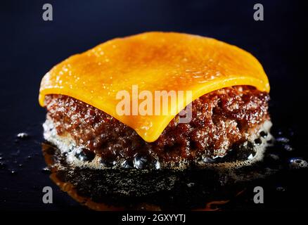 Leckerer Cheeseburger mit Rinderpastete, die in Nahaufnahme auf einem Grill auf dem geschmolzenen Käse brutzeln, und saftiges Fleisch auf dunklem Hintergrund für die Menüwerbung Stockfoto