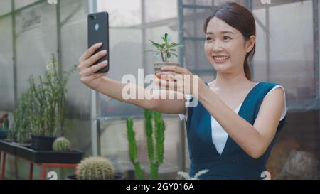Happy Asian schöne junge Frau, die Selfie-Foto von kleinen Baum auf Topf von Smartphone zum Verkauf im Garten in der Nähe von Gewächshaus, weibliche Gärtner Video cal Stockfoto